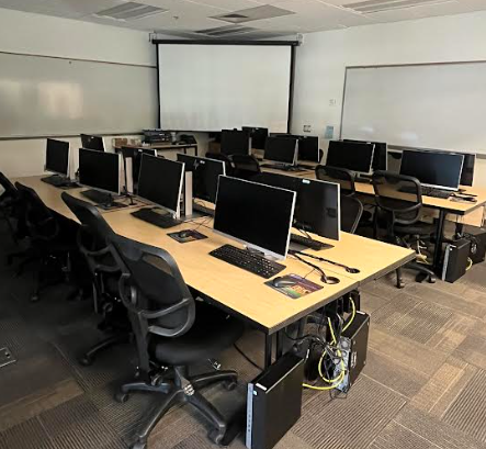 A classroom with two long tables. On them are rows of computers. A projector screen and two whiteboards cover the far wall.