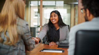 A smiling interviewee shakes the hand of one of two interviewers facing her.