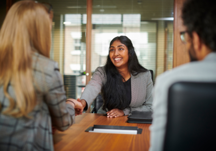 A smiling interviewee shakes the hand of one of two interviewers facing her.