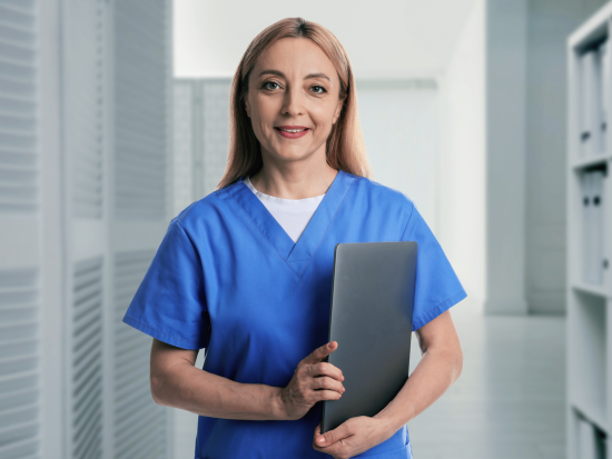 A medical administrative professional wearing blue scrubs stands in a bright healthcare hallway, holding a tablet and smiling confidently toward the camera.