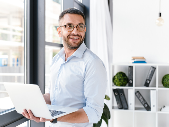 Professional man holding a laptop and smiling while standing near a window in a bright, modern office.
