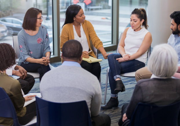 A group therapy session featuring men and women sitting in a circle listening to a woman speak.