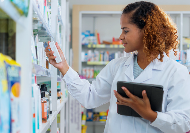 Female pharmacy technician in a pharmacy setting holding a tablet and looking at a medicine box taken from a wall of medical supplies.